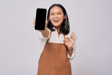 Smiling young Asian woman barista barman employee wearing brown apron working in coffee shop showing mobile phone blank screen mockup and okay sign isolated on white background. Small business startup