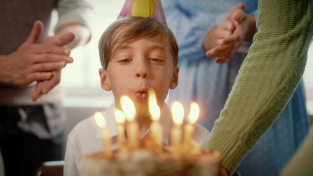 Cute seven-years boy blowing candles on cake, celebrating birthday, portrait. Slow motion, parents and family congratulating little child and flapping hands, happy childhood moment, growing up process