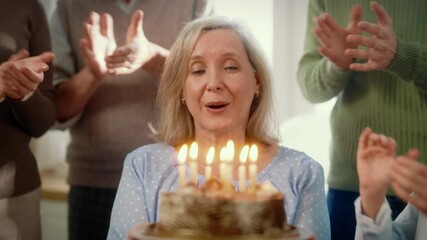 Charming elderly woman blowing candles on traditional birthday cake, slow motion . Happy old woman celebrating her anniversary with family, change of age and aging process, retiree female person
