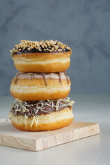 three medium sized donuts topped with chocolate sprinkles, tiramisu nuts and cheese, stacked together, on a wooden board photographed with studio light