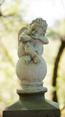 angel statue in the park. little boy with wings. cupid statue in spring park. figurine for design, marble sad angel sitting on a ball. blurred natural background.