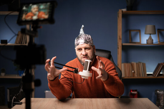 Strange stuiped bearded man vlogger in foil hat speaking with his subscribers on camera, He recording live stream at home. Fake news conspiracy theory