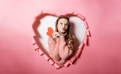 Charming young woman in pink sweater with long hair smiling happily holding red heart decoration. Perfect combination of innocence, love and spirit valentines day, celebration youthful joy and fashion