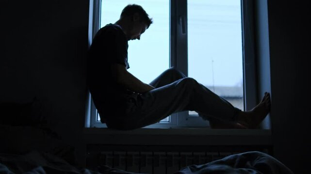 Silhouette of a man sitting on a windowsill, holding his head, showing sadness and emotional struggle in a dimly lit room. A man in depression and existential crisis