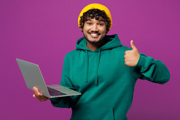 Young smiling happy Indian IT man wear green hoody yellow hat casual clothes hold use work on...