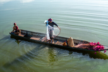 Obraz premium Fisherman casting his net at sunrise. Silhouette Asian fisherman on wooden boat casting a net in the Lake. Vietnamese Fisherman throwing net.