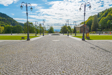 Big square with cobblestone floor at the Oropa Shrine, located close to the city of Biella, Italy. Elegant street lamps at both sides, with green garden. Blue sky with white clouds on the background