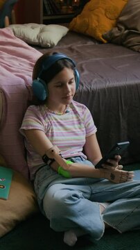 Vertical view of teenage girl with upper limb prosthesis surfing internet on smartphone and listening to music with headphones while sitting on carpet in bedroom