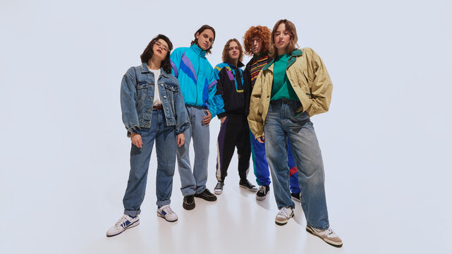 Diverse group of young adults dressed in classic 90s-inspired outfits, including oversized denim, colorful windbreakers, and retro sneakers, standing against white background. Concept of 90s fashion