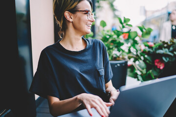 Cheerful hipster girl looking aside and smiling in break of course language online via laptop,...
