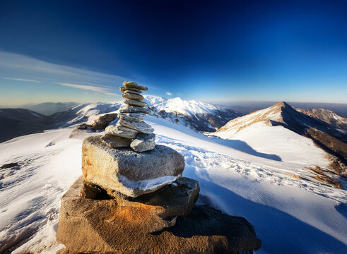 A cairn of balanced stones sits atop a snow-covered mountain peak, overlooking a vast, sunlit panorama of snow-capped peaks under a vibrant blue sky.  A sense of serenity and accomplishment is evoked.