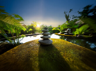 Serene sunrise illuminates a carefully balanced stack of grey stones nestled on moss-covered rock beside tranquil water, lush tropical foliage surrounds.