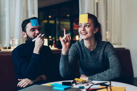 Couple of positive teenage friends gathered together in and playing who am I game while spending time at cafeteria, pretty young hipster girl trying to guess what is written on her sticky note