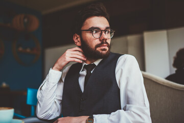 Thoughtful hipster guy in spectacles for vision correction sitting at cafeteria table with coffee cup and pondering on leisure, young pensive man in trendy apparel waiting friend at cafe interior
