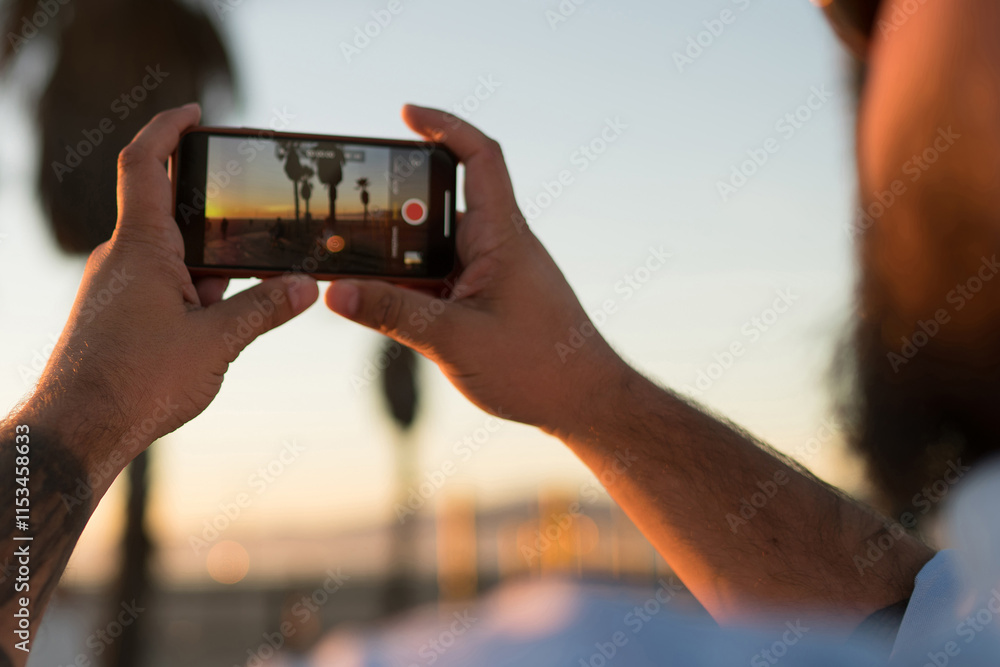 Poster back view of young man using smartphone camera for taking pictures of sunset and share in social net