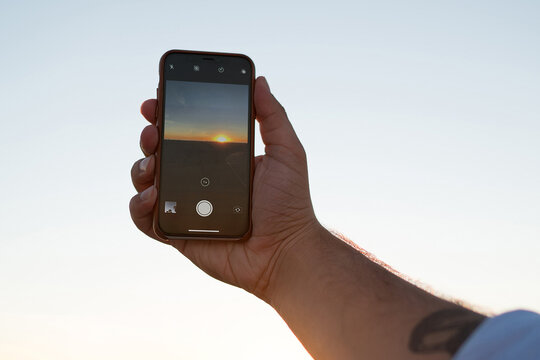 Cropped image of man's hand holding smartphone shooting video of evening sky and sundown, male using modern mobile phone camera taking photo of golden sunset  and nature during summer evening - Powered by Adobe