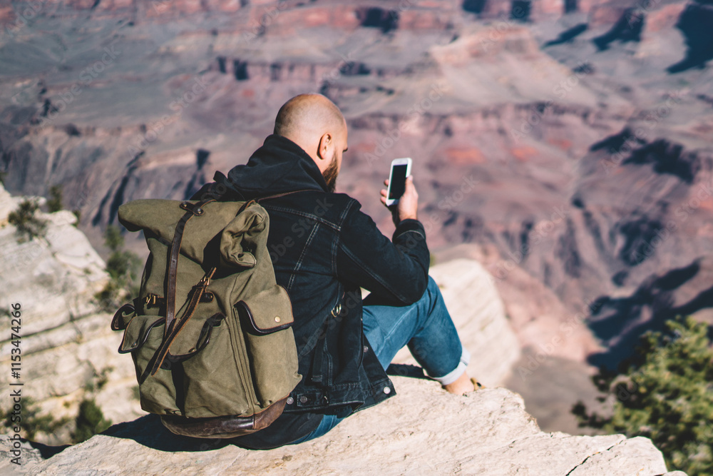 Poster hipster guy with rucksack sitting on high mountain cliff using smartphone for reading text message i
