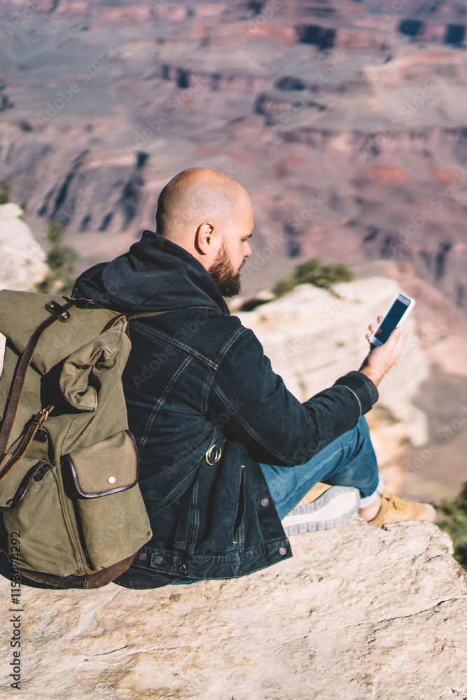 Poster hipster guy with rucksack sitting on high mountain cliff using smartphone for reading text message i