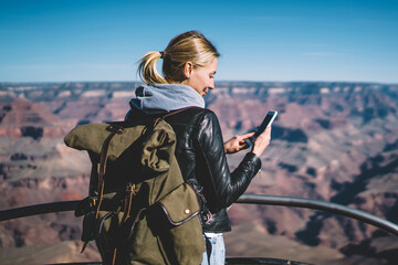 Smiling young hipster girl wanderer with backpack checking good mobile connection hiking on...