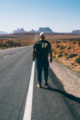 Fototapeta premium Back view of man with backpack walking on asphalt road in National park in southwest of USA, male traveler wanderlust exploring America going in trip to Monument valley while hitchhiking on the way