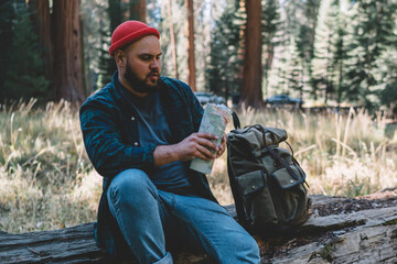 Male traveler wanderlust rest while trekking in wood checking route on map discovering nature environment, hipster guy sitting on old tree with backpack exploring wild forest during journey in USA
