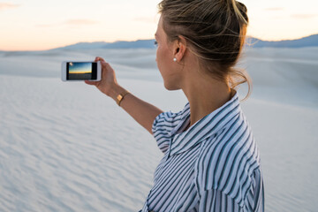 Hipster girl holding mobile phone making picture visiting famous american landmark White sands Park at sunset, back view of young woman using smartphone camera taking photos of scenic nature landscape