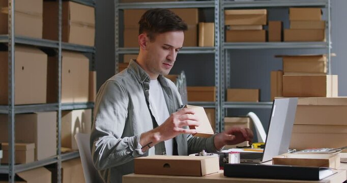 Man is using a laptop in a warehouse or postal office, conducting inventory of packages and processing orders. He is focused on managing storage and preparing packages for shipment.