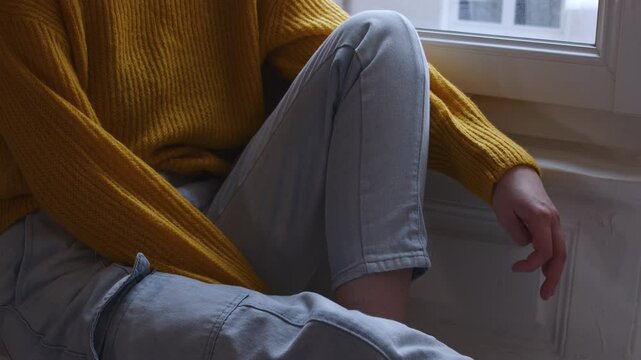 Young woman sits by window fiddling with her fingers stressed thinking