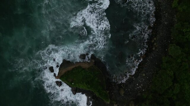 Aerial slow-motion of a green hill with tranquil ocean waves on Menganti Beach in Kebumen, Indonesia