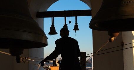 Bell ringer skillfully pulling ropes high in Orthodox bell tower at sunset, filling sky with...