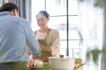 A woman prepares fresh vegetables in a cozy kitchen, while a man assists in the background. The scene emphasizes a warm and healthy lifestyle with a focus on fresh produce and home cooking.