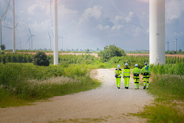 A team of engineers in high-visibility safety gear stand together at a wind farm, conducting...