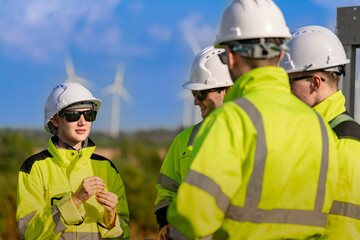 A team of engineers in high visibility safety gear stand together at a wind farm, conducting inspections and discussing renewable energy projects. Wind turbines surround them under a clear sky.