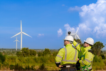 A team of engineers in high visibility safety gear stand together at a wind farm, conducting...