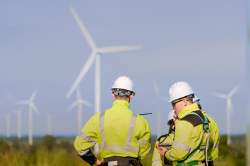 A team of engineers in high visibility safety gear stand together at a wind farm, conducting...