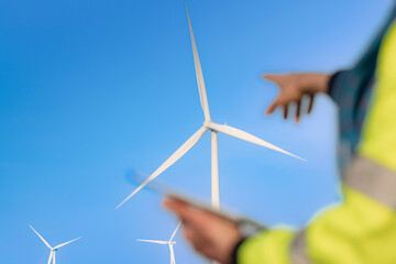 A blurred figure holding a tablet observes large wind turbines in the background under a clear blue...