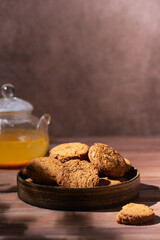 Oatmeal cookies in a plate with sea buckthorn tea in a teapot on a wooden table. Breakfast concept. Hard light. Vertical orientation.