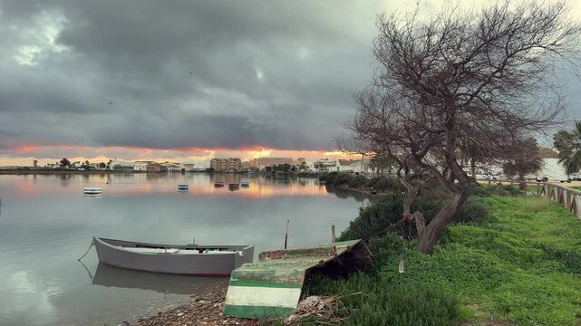 Tranquil scene of a fishing boat moored on the barbate river in barbate, andalusia, spain, at dawn under a dramatic cloudy sky, with reflections of the town and colorful sunrise on the calm water
