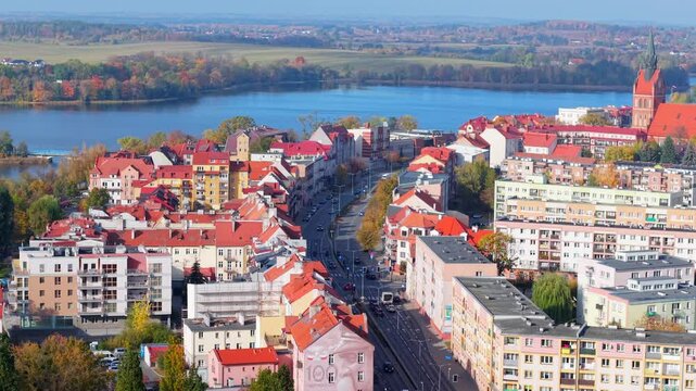 Drone follows Wojska Polskiego street in Elk, Poland, lake along fields, and orange roof city landmarks with church of the sacred heart of Jesus