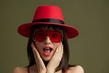 Woman, red hat, sunglasses a woman in a vibrant red hat and stylish sunglasses stands against a green background, her mouth open in shock and surprise, capturing a dramatic and unexpected moment