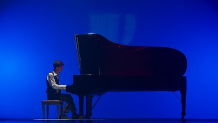 Pianist sitting and playing on the black grand piano on dark concert stage under bright spotlight, isolated on blue studio background. Artist performing in concert hall. © kinomaster