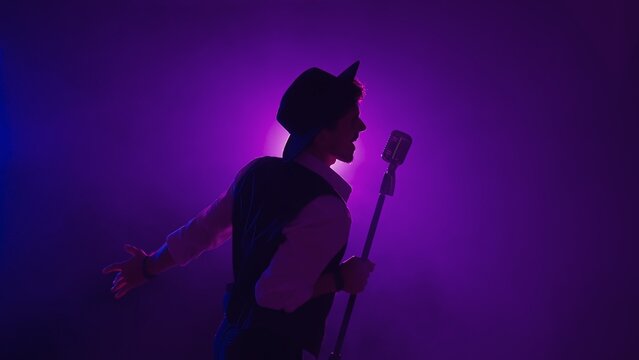 Man musician in stylish hat standing on dark concert stage against purple spotlight and singing in retro microphone, isolated on black studio background.