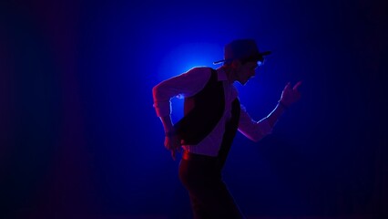 Man musician in stylish hat dancing on dark concert stage against blue spotlight and singing looking at camera, isolated on black studio background.
