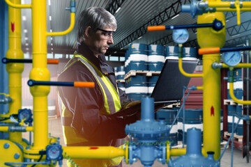 Man technologist at fuel company. Gas pipes near engineer with laptop. Fuel barrels behind technologist. Fuel station contractor inside industrial building. Man in yellow vest works in enterprise