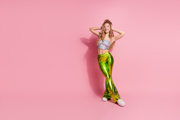 Trendy young woman in shimmering green pants poses confidently against a vibrant pink background, embodying style and glamour with a radiant smile and relaxed hands-on-head gesture.