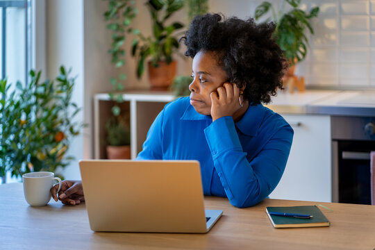Melancholic African American woman feeling apathy, stress life. Frustrated black female deep in bad thought, sadly looking out window avoiding work, sits at desk with open laptop and untouched coffee