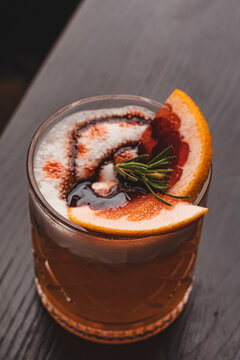 C&oacute;ctel sour de autor en vaso corto labrado con abundante espuma blanca, piel de naranja y romero. Trago gourmet sobre mesa de m&aacute;rmol en un bar.