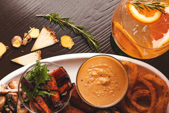 Vista cenital de tabla de picoteo con aros de cebolla, bastones de vegetales asados, champi&ntilde;ones y salsa dip junto a un c&oacute;ctel refrescante en mesa de madera oscura.