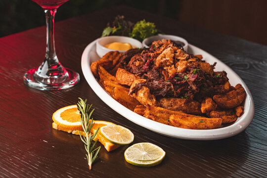 Chorrillana tradicional chilena con papas fritas r&uacute;sticas, carne mechada y salsas en un bar. Plato para compartir con c&oacute;ctel en mesa de madera.