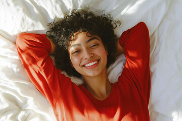 A young woman with curly hair smiles contentedly while lying on a white bed in a red longsleeved shirt, arms behind her head.